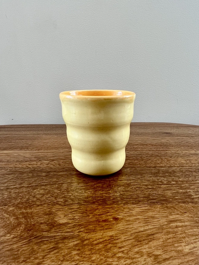 ceramic wavy tumbler with butter yellow glaze on the outside and orange on the inside. the tumbler holds 250ml of liquid and sits on a wooden table