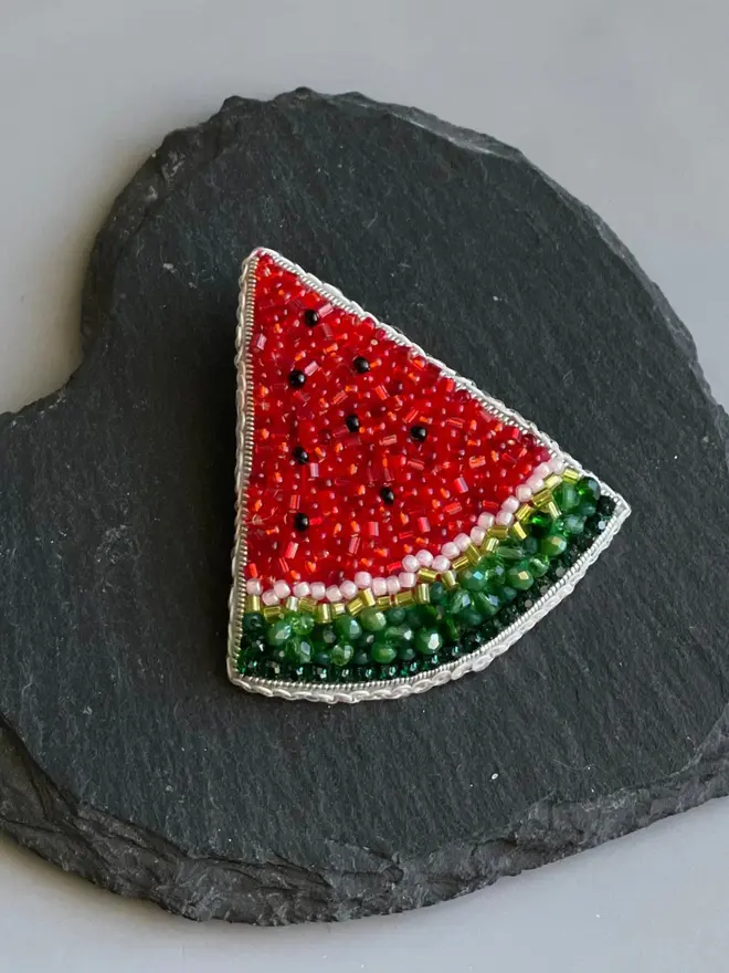 Watermelon Beaded Brooch displayed on a black slate in the shape of a heart, against a grey background. 