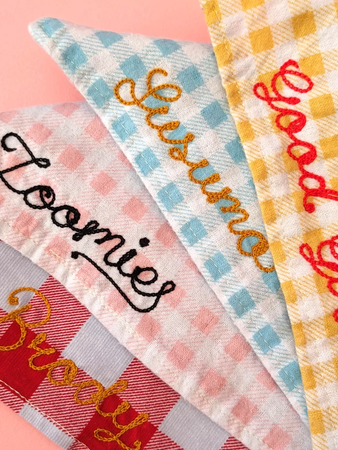 The complete range of dog bandana colour-ways stacked together, each personalised in a different colour and word, on a pink background