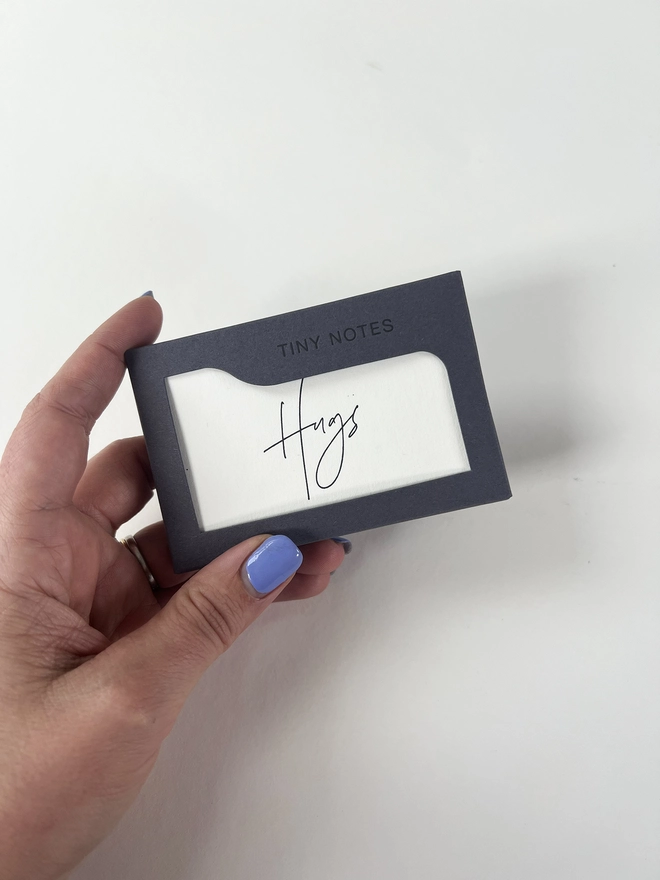 A dark grey box of modern calligraphy style flat tiny notes and envelopes is held in a woman's hand to show scale. The box say's Tiny Notes on the front of the box.