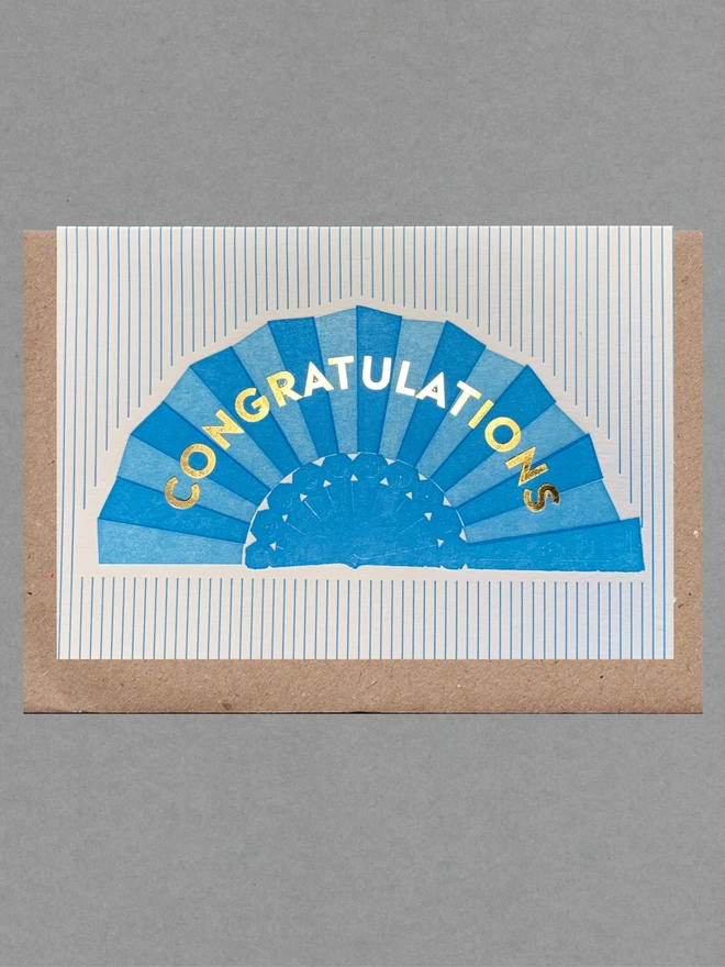 Blue and white striped card with a blue fan on it and gold text reading 'Congratulations' with a brown envelope behind