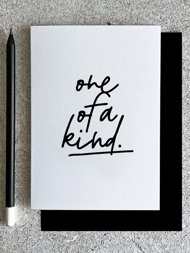 Black and white typographic card saying 'One of a Kind' with pencil on mottled background desk