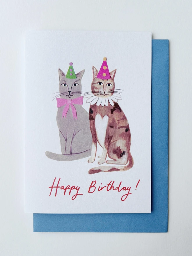 A birthday card featuring two cats wearing party hats. The message on the front of the card reads "Happy Birthday!" in a handwritten cursive script 