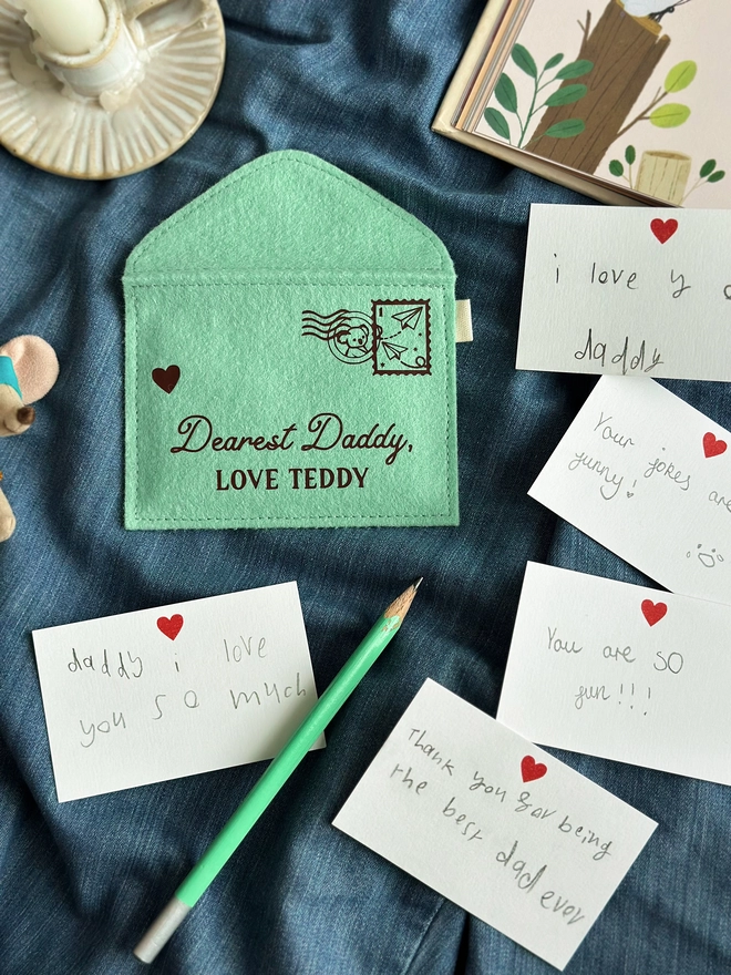 A handmade green felt envelope with the personalisation 'Dearest Daddy, love Maude' and a paper aeroplane stamp with postmark, lays on dark blue fabric. Notes to Daddy are written on the included heart notecards.