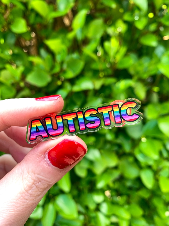 image shows a hand holding acrylic badge in the shape of the word 'autistic'. the letters are filled with horizontal rainbow stripes.