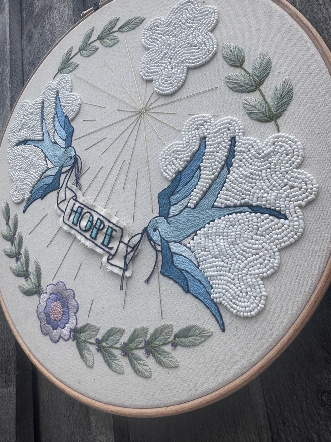 An embroidery hoop containing embroidered swallows beaded clouds and a banner saying hope