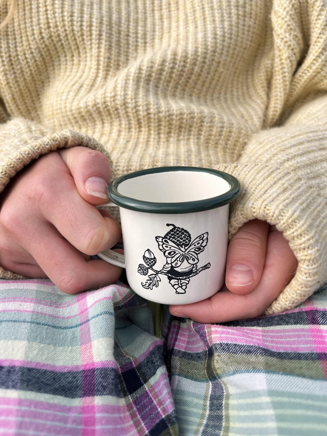 a mini cream enamel mug with a dark green rim featuring an etching of a pixie sat on a twig wearing an acorn hat