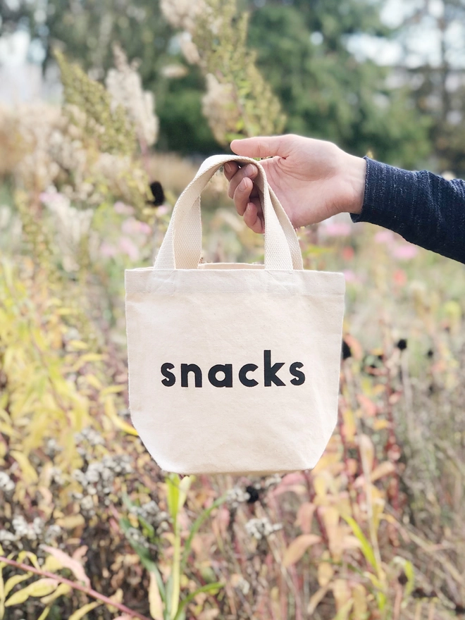 a hand holding out a mini kid's size tote bag with the word snacks on it