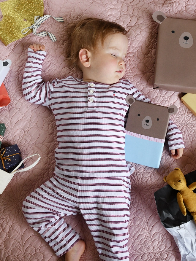 A baby wearing red and white striped pyjamas is asleep on a pink quilt with three gifts wrapped up as teddy bears.