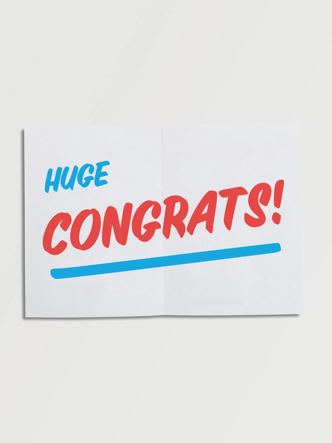 Unfolded A5 piece of paper that says BIG HUGE CONGRATS! in red fun slanted typography on an angle with a blue underline