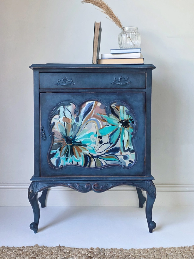 A french style bedside cabinet or drinks cabinet with beautiful carving especially on the legs. The frame has been painted a rich navy blue to contrast against a bespoke handpainted glass panel in the centre featuring daisies in turquoise, pink, white, and rusty orange. It is centre stage sitting in front of a rustic rattan rug, and sitting on top, vintage books, a reeded glass vase and pampas grass.