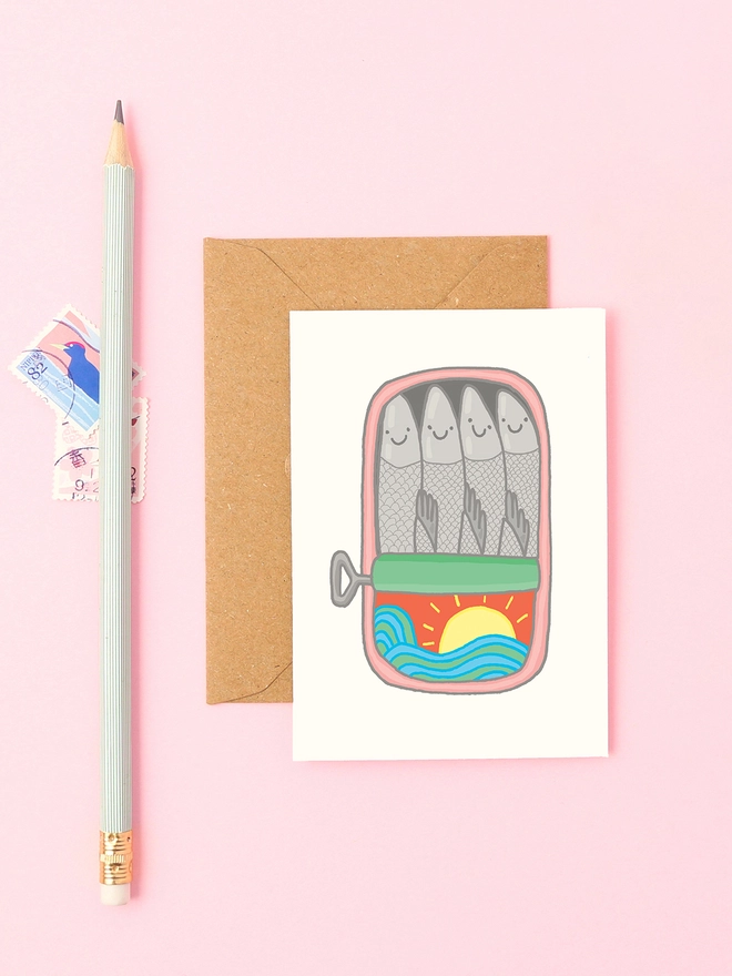 Mini sardines greeting card styled with kraft envelope, pastel pencil and vintage stamps on a pink background. Features a hand-drawn tin of happy sardines with sun and sea illustration.