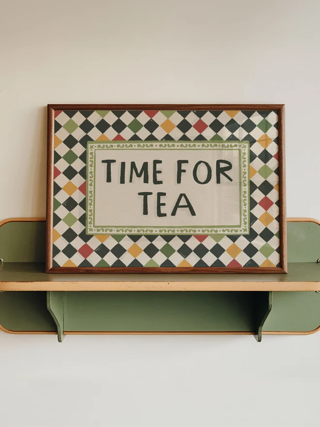 'Time for tea' checkerboard art prints on a wooden shelf on a white and green wall. 