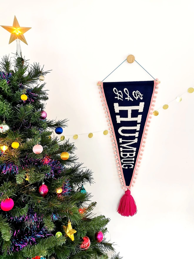 A navy knitted pennant flag wall hanging with blush pink pom pom trim hangs on a wall next to a colourful Christmas tree hung with retro glass baubles and a lit up star on top. The words ‘Bah Humbug’ are written across the banner in white glittery retro writing and an oversized raspberry pink tassel hangs from the point of the banner.