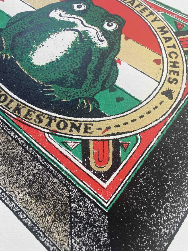  An image of a limited edition A2 screenprint. The print depicts a retro-style matchbox illustration with a central image of a green frog with a white belly, set against a background of horizontal red, white, and green stripes. The frog is framed by a gold-bordered oval. Text at the top of the oval reads "PRINCE CHARMING SAFETY MATCHES," and at the bottom, "MADE IN FOLKESTONE." The matchbox border is red with ornate details and a green trim. The match tips on the left are a textured gold. Below the main image, the print is signed and numbered 5/11. The entire image has a grainy texture, characteristic of a vintage screenprint.