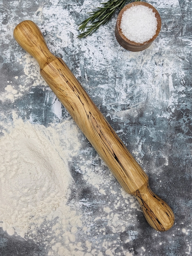 A stunning hand made rolling pin in Spalted Beech by Something From The Turnery. The rolling pin sits on an art house kitchen top amongst flour, rosemary and a wooden salt pot, prepped to bake some delicious bread! Made from Spalted Beech, the rolling pin features two traditional shaped handles amongst it’s own , never repeated natural grain patterns, travelling through golden browns, tans and stunning black lines to outline the changes in the timber. 
