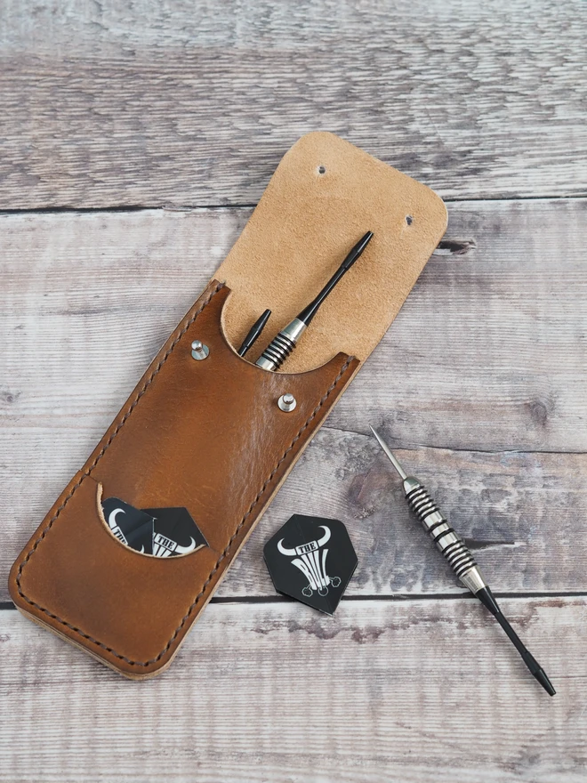 Personalised leather darts pouch. A handmade leather sleeve with 2 darts inside the case and one waiting to be put inside. The same amount of flights sit in a slim external front pocket.