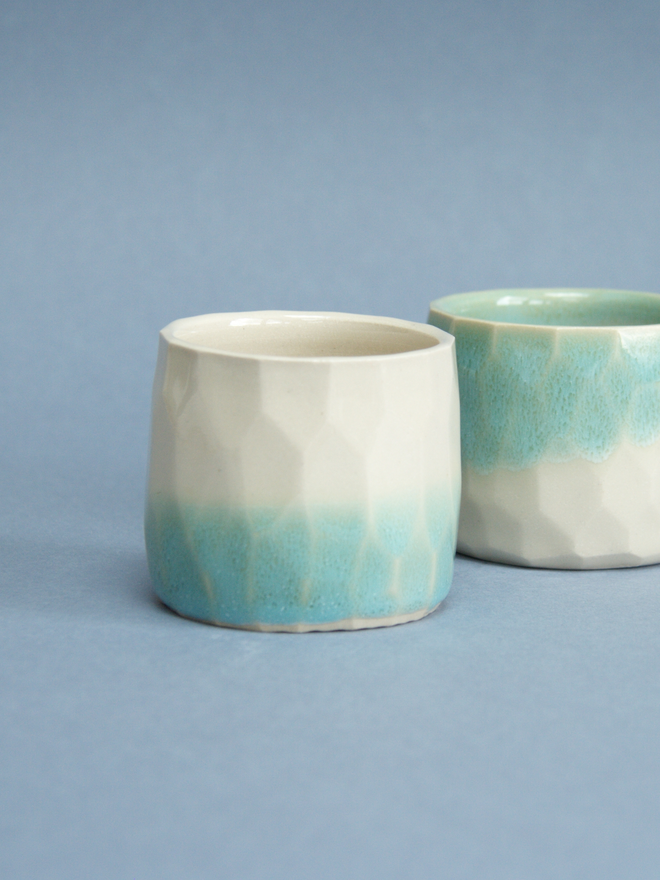 Two straight sided ceramic cups next to eachother.  One has a turquoise top half fading into a glossy cream bottom half.  The other cup as the opposite, with the top half being glossy cream and the bottom half being turquoise.The surface of the cups have a hexagonal texture.
