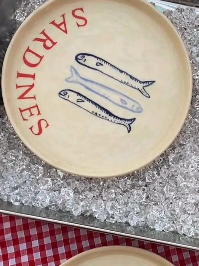 'Sardines' Red Wording Ceramic Side Plate With 3 Fish, a ceramic side plate sitting on ice with a fish design. 
