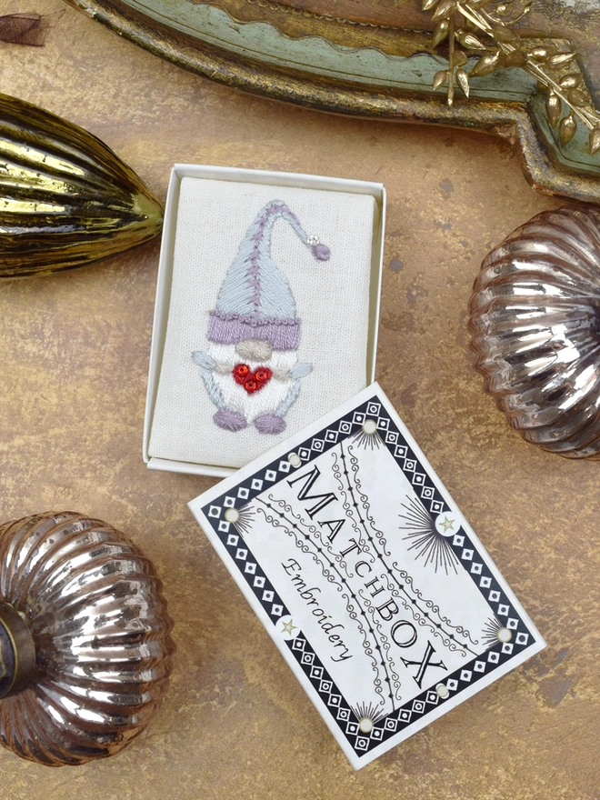 The embroidered Gonk, inside a Matchbox displayed on a gold background with glass baubles.