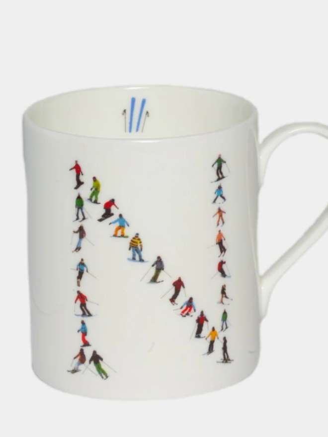 A simple white mug illustrated with an ’N’ made of small, multicoloured skiers. The mug is set against a plain, white background.