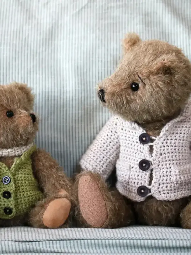 Brown teddy bear wearing a cream knitted cardigan sitting among a group of other teddy bears.