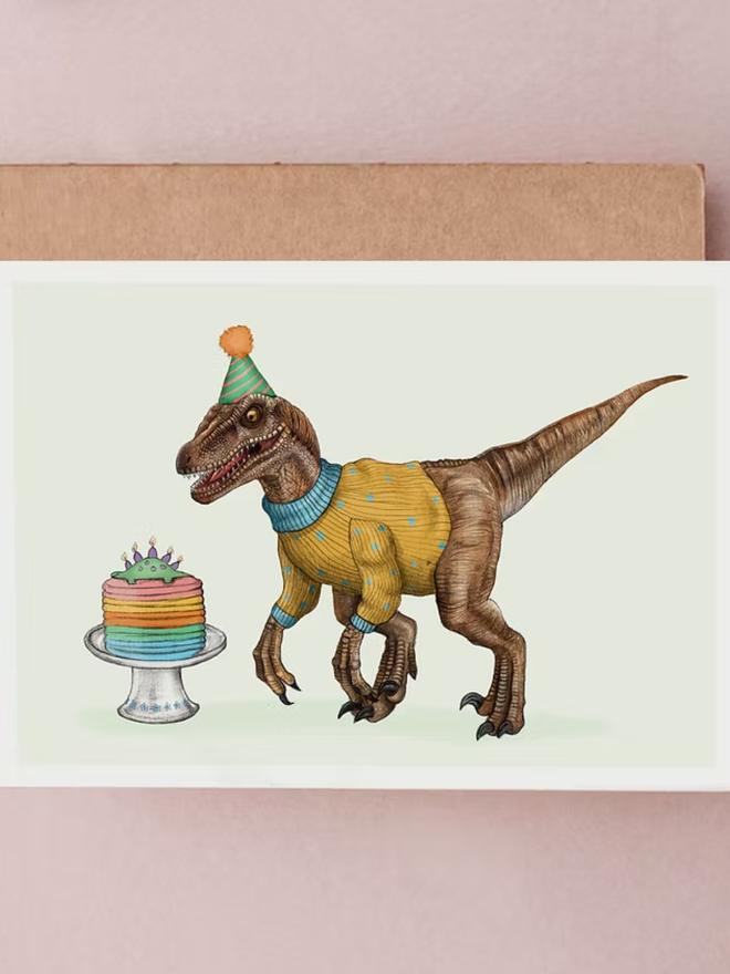 illustrated velociraptor dinosaur birthday card. a drawing of a dinosaur wearing a jumper and party hat, with a birthday cake.