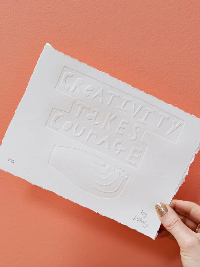 'Creativity Takes Courage' embossing on white paper, signed by the artist and held up against a coral wall by a woman's hand