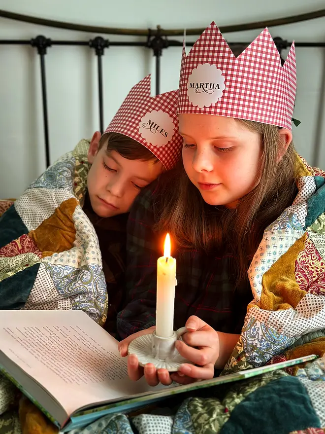 Two children wearing red gingham personalised Christmas crowns are wrapped in a cosy patchwork quilt reading a book together by candlelight.