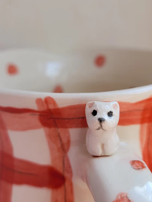 a seated white dog on the top of a handle on a ceramic red check cup