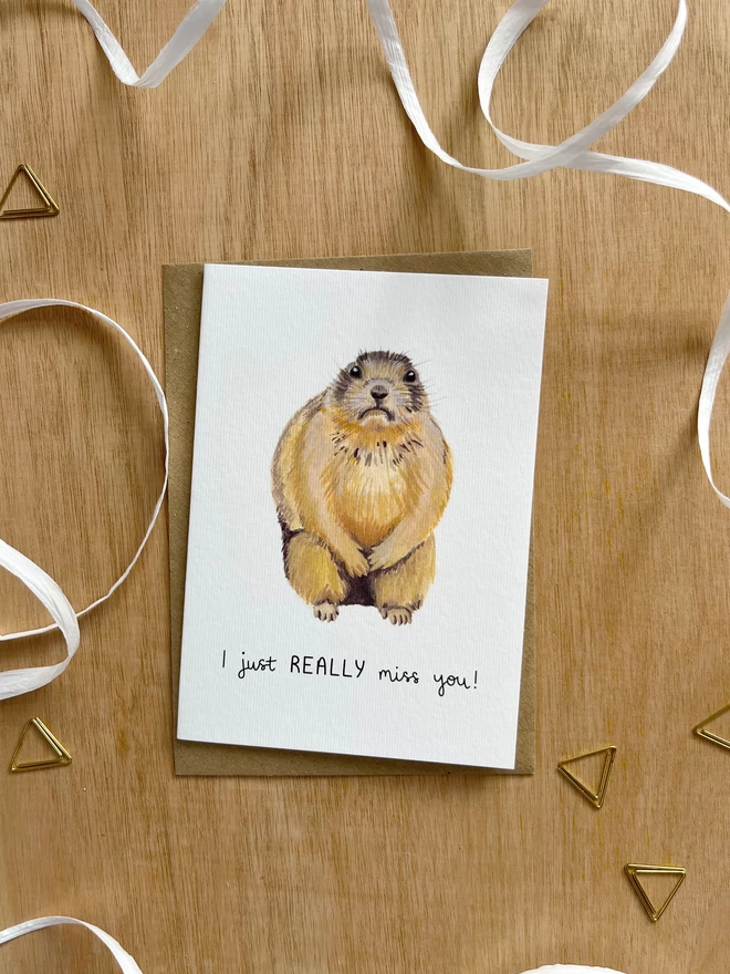 a greetings card with a white background featuring an illustration of a sad looking prairie dog with the phrase “I just really miss you”