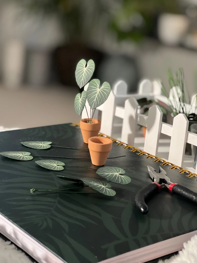 A miniature replica Philodendron Gloriosum paper plant ornament in a terracotta pot being crafted with paper leaves and a pair of pliers in shot