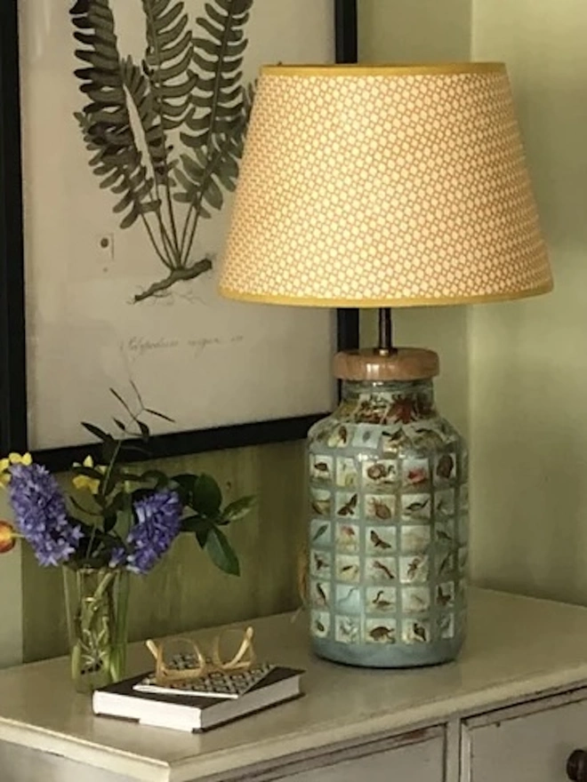 Retro Drum Lampshade. A peach diamond lattice lampshade with a mustard trim on a decoupage glass lamp base, styled on a painted chest of drawers beside spring flowers. The block-printed shade complements a botanical print for a relaxed country home feel.