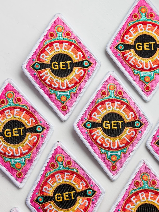 Seven Rebels Get Results patches are laid out on a white background. The patch is a diamond shape embroidered patch with “Rebels Get Results” at the centre. The design features pink, white, red, yellow and black colours and has a white border. 