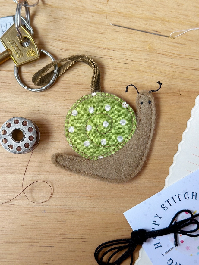 A homemade felt snail keyring with a brown body and a green spotty shell lays on a wooden desk.
