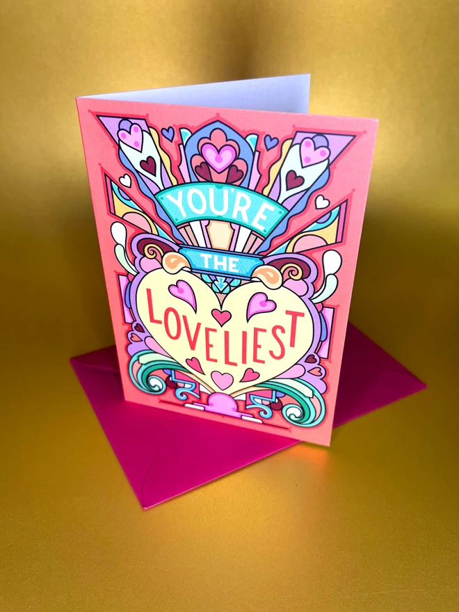 A greetings card with the phrase “You’re the Loveliest” surrounded by an abstract multi-coloured design and lots of hearts, sits on a red envelope in front of a gold backdrop.