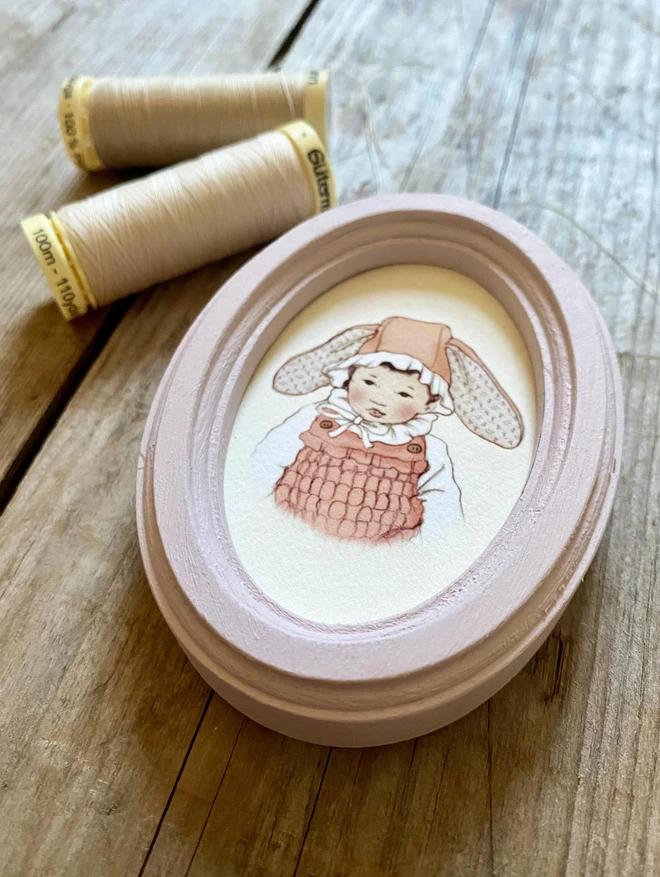 a photograph of a miniature personalised easter bonnet portrait framed in a dusty pink wooden oval frame japanese baby toddler