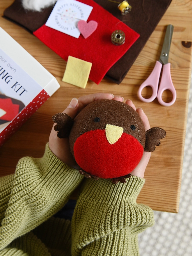 A small round robin plush toy is being held in two hands above a wooden desk where craft kit contents are laid out.