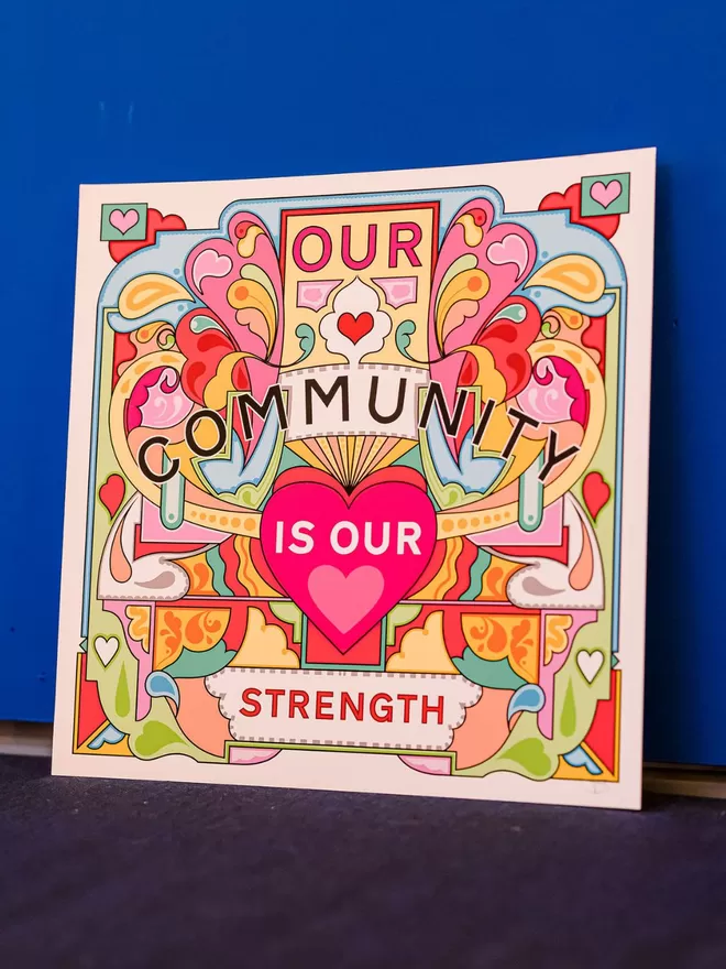 Our Community is Our Strength is written over this bold, symmetrical illustration of yellows, greens and pinks. The print sits on a dark carpet and is resting against a dark blue wall. 