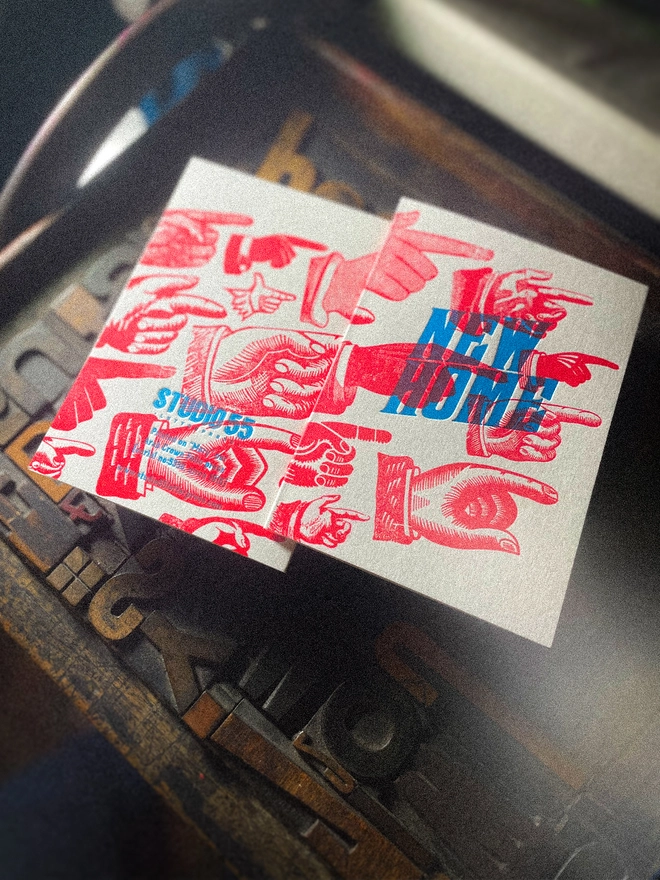 A double-sided, hand-printed letterpress artisan moving house announcement card with bold blue “New Home” typography and fluorescent red pointing hand illustrations (fists) on thick premium white card.