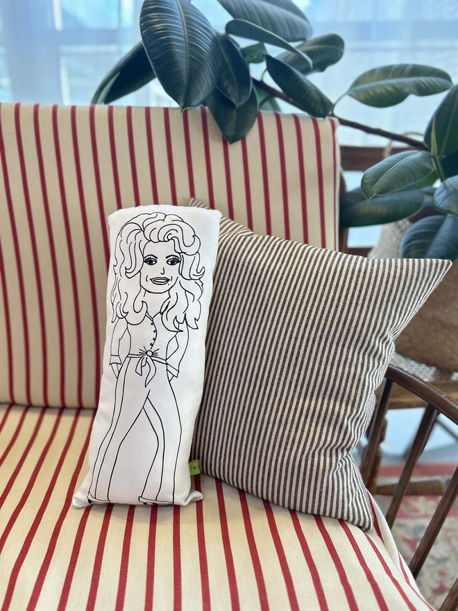 Black and white illustrated screen printed cushion of Dolly Parton. The cushion sits on a striped sofa.