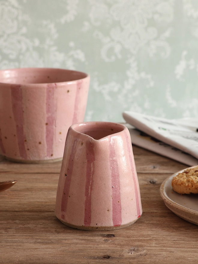 Handmade Ceramic Stoneware Small Jug in Pink Stripe sat on a wooden surface. A mug in the same style and a newspaper sit behind a mug, while a plate of cookies can be seen to the right. 