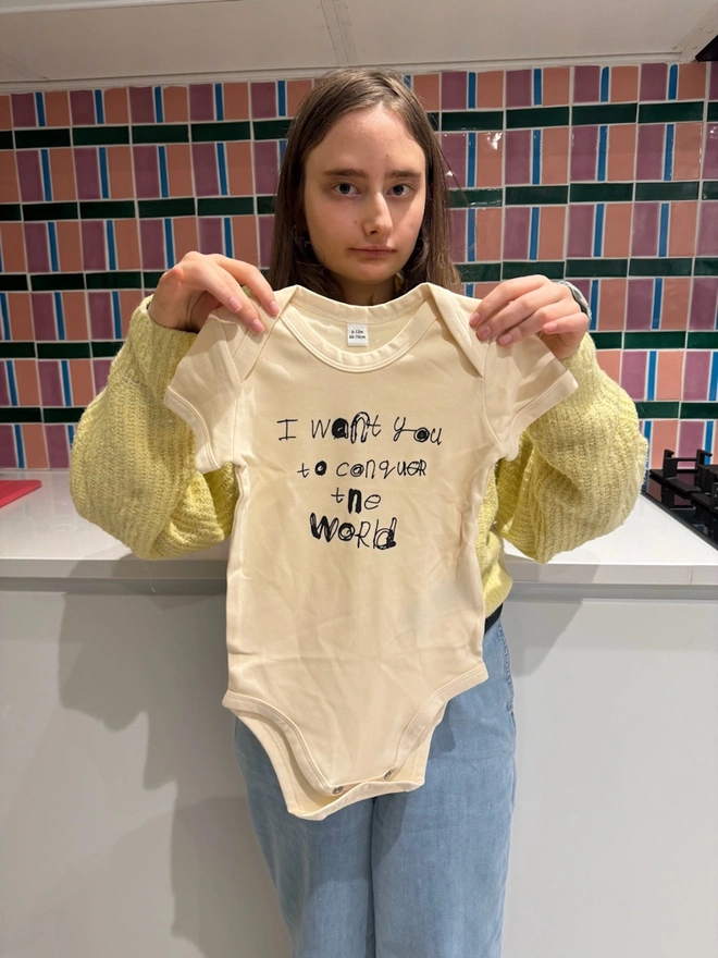 Vanilla off white colour baby grow with Black text held up by Piper