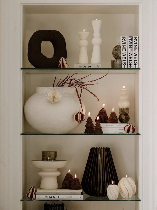 Brown Christmas Tree Warm Candles (Set Of 2). Glass shelves styled with lit chocolate brown Christmas tree candles, cream ribbed totem candles and swirled pillars, alongside a textured white vase, paper baubles and stacked books. The sculptural candles add warmth and a modern festive touch to a styled alcove.