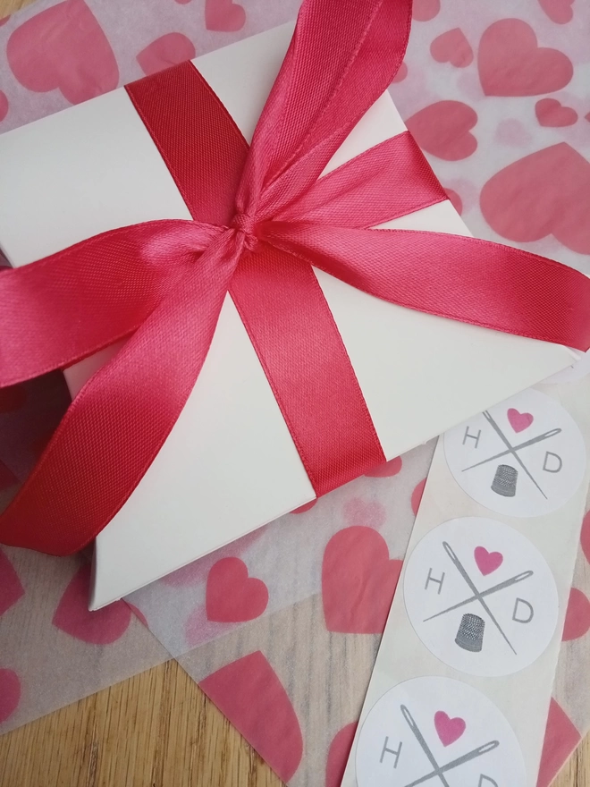 A white pillow box tied with a pink ribbon, sitting on top of pink and white heart print tissue paper with some round sewing logo stickers