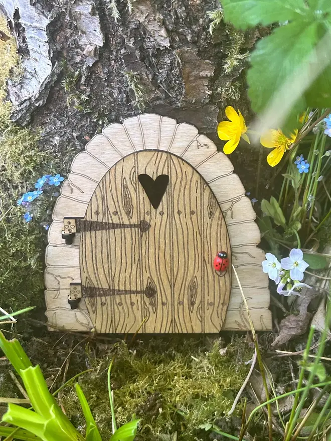 Wooden fairy door for kids with engraved woodgrain detail, heart-shaped window, and decorative hinges, displayed at the base of a tree surrounded by moss and wildflowers.