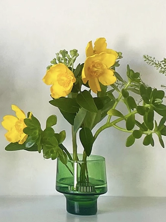 Small Green Flower Arranging Items (Set Of 2), a green glass vase sitting on a plain surface against a white backdrop. It is filled by a bundle of flowers grouped together by a copper frog. 