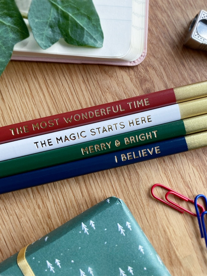 Four festive coloured pencils with gold writing along each one lay on a wooden desk beside various stationery items.