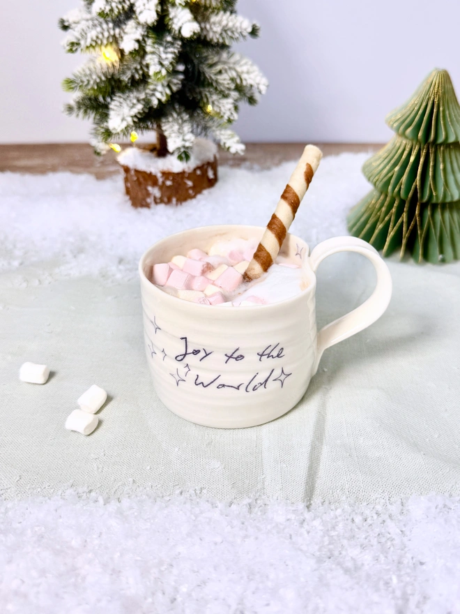 cream hand thrown porcelain mug with the words 'Joy to the World' printed onto the side with pretty line drawn stars. Sitting on a tabletop with snow, a mini lit Christmas tree and filled with a luxurious hot chocolate complete with marshmallows, thick cream and a chocolate straw