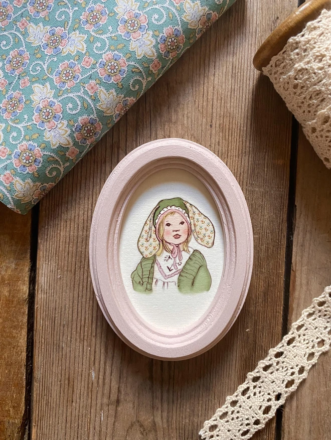 a photograph of a tiny personalised portrait of a sweet little girl with blonde hair wearing an easter bonnet with bunny ears drawn in a vintage children's book style. Framed in a dusty pink tiny hand made oval wooden frame. photographed on a wooden bench with ribbon and floral fabric